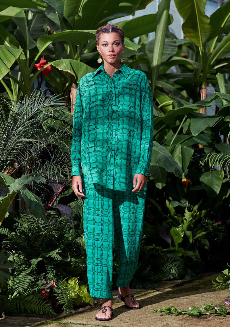 A woman models a patterned green shirt and pants surrounded by tropical foliage in an eye-level, medium shot, showcasing a fashion editorial style.