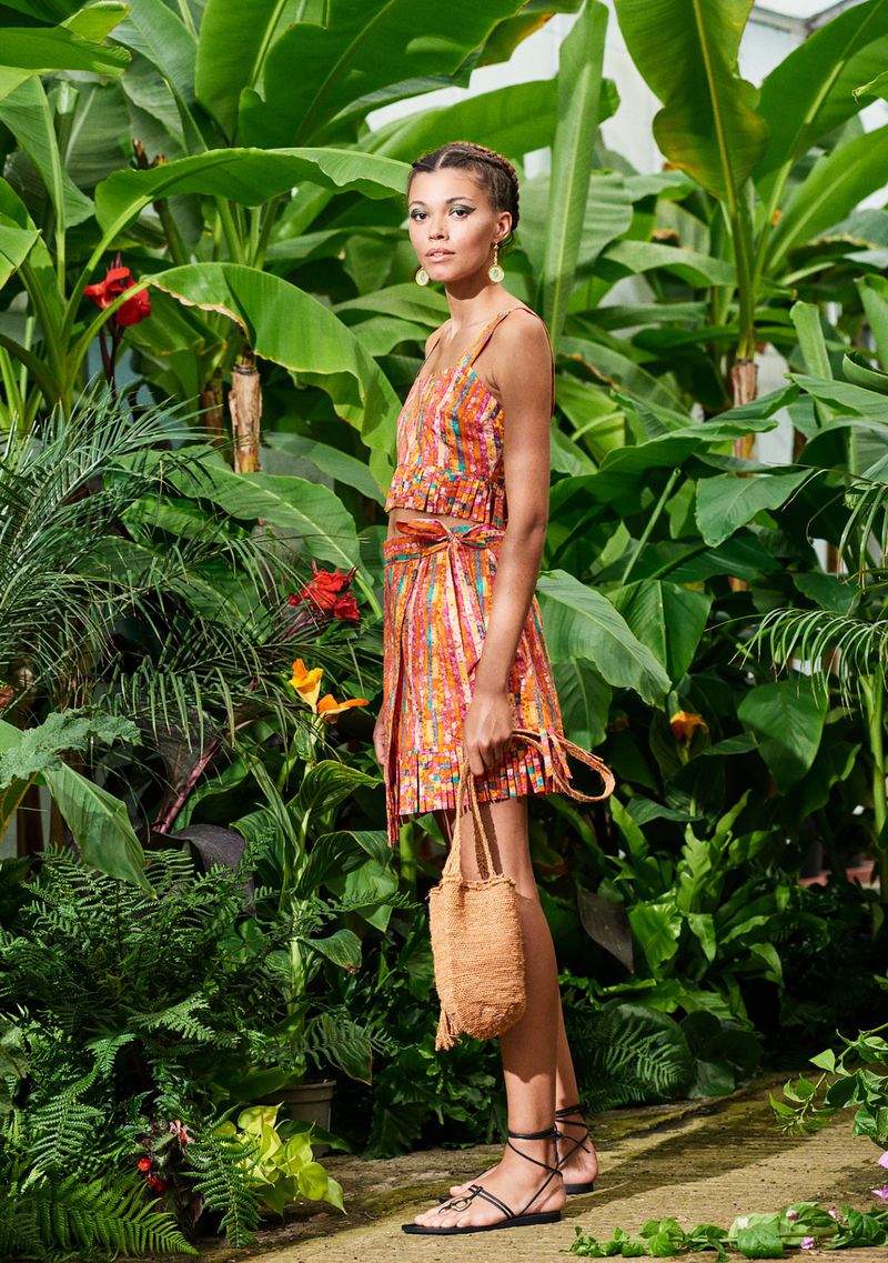 An adult woman poses in a two-piece orange print dress, accessorized with a straw bag and sandals, surrounded by tropical foliage.