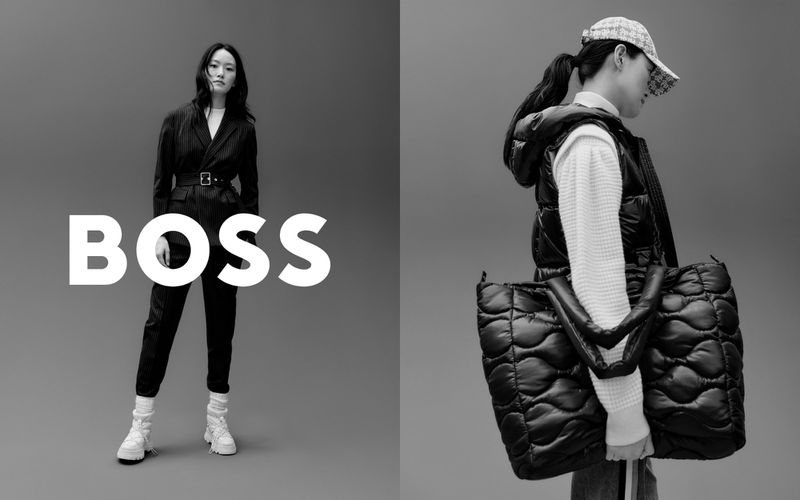 Black and white fashion shot of two Asian women modeling BOSS-branded clothing and accessories against a gray studio backdrop.