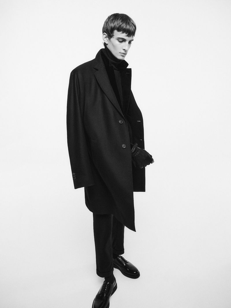 A full shot in black and white of a male model wearing a dark overcoat, turtleneck, gloves, trousers and leather shoes against a white background.