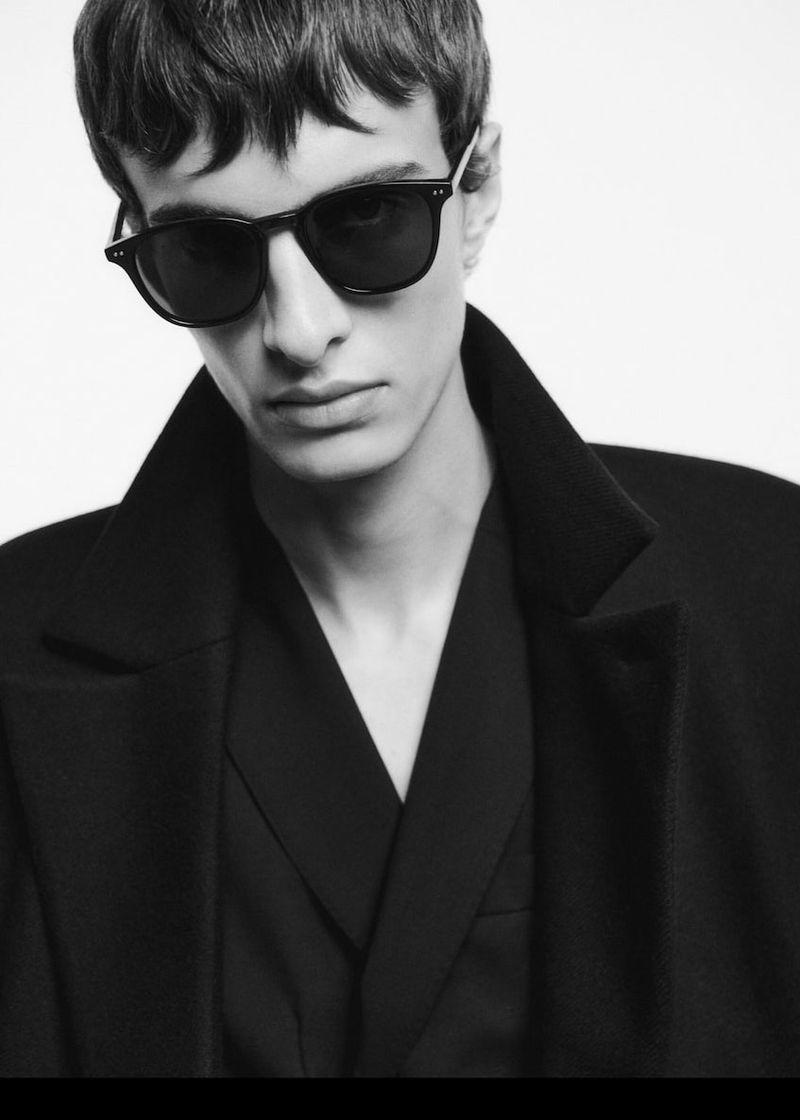 Headshot of adult male wearing sunglasses and black overcoat in a studio setting with soft, diffused lighting on a white backdrop.