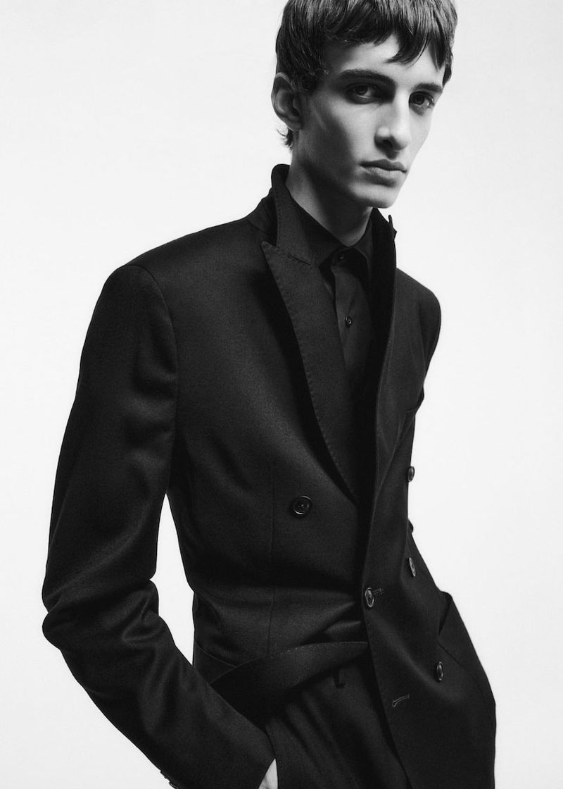 A young adult male fashion model poses in a sleek black suit against a clean white backdrop, showcasing modern tailoring.