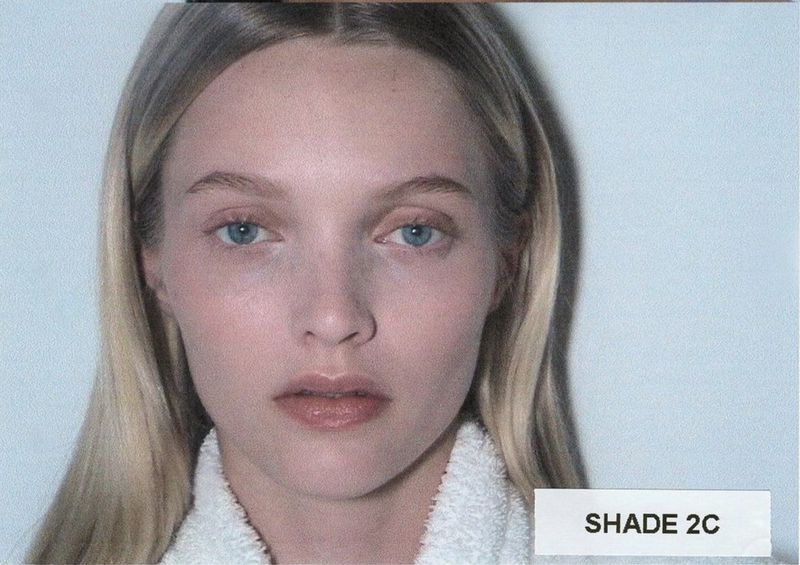 Close-up of a blonde woman with light skin and blue eyes, wearing a white towel around her shoulders, against a pale blue background.