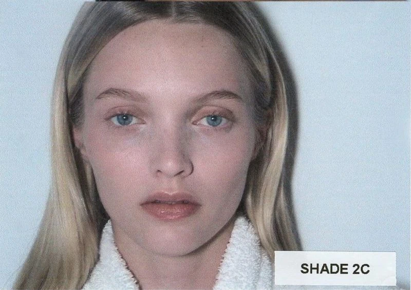 Close-up of a blonde woman with light skin and blue eyes, wearing a white towel around her shoulders, against a pale blue background.