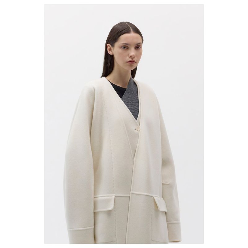 A woman in a neutral-toned, oversized coat in a studio setting, captured in a medium shot with soft lighting.