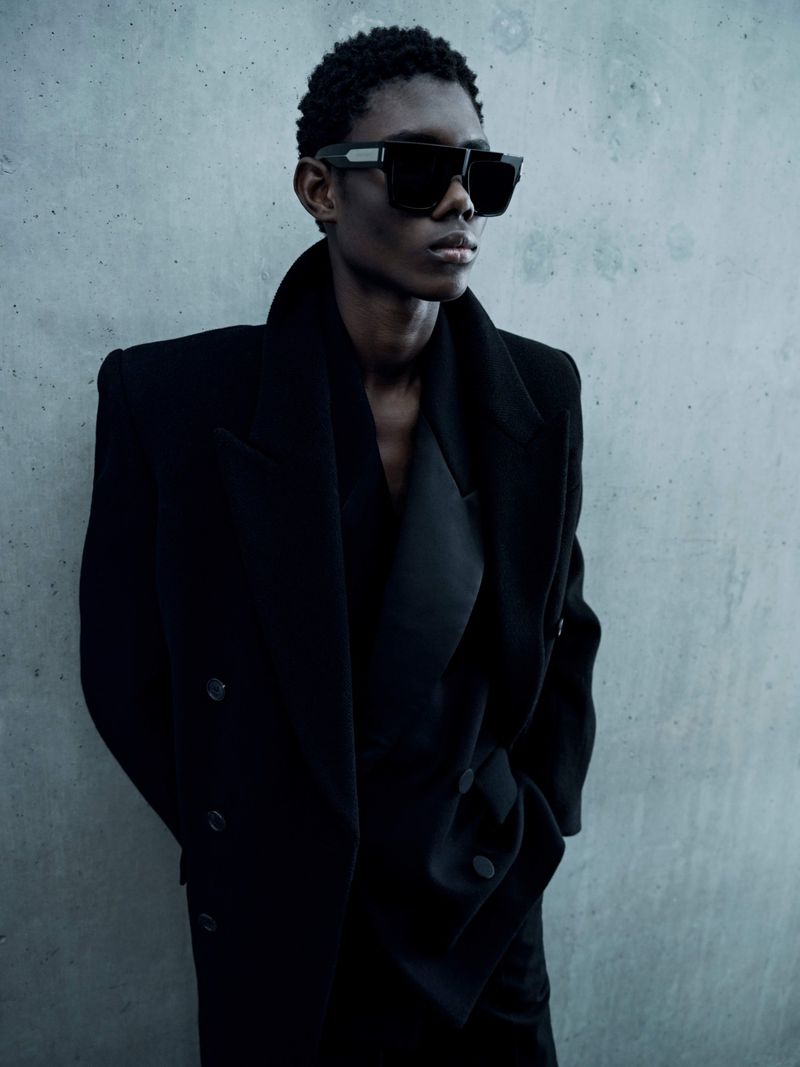 A stylish Black male model poses in a black coat and sunglasses against a textured gray backdrop in a fashion-editorial portrait.
