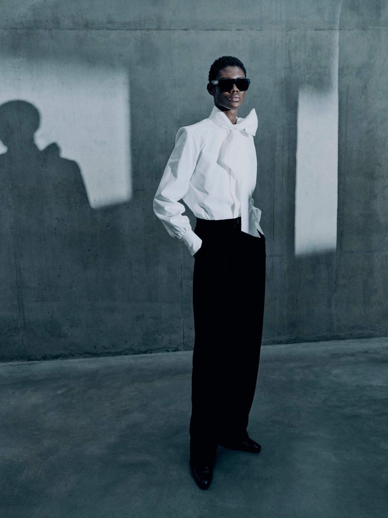 Fashionable Black male model poses in a white dress shirt and black trousers against a textured gray wall with window shadow patterns.