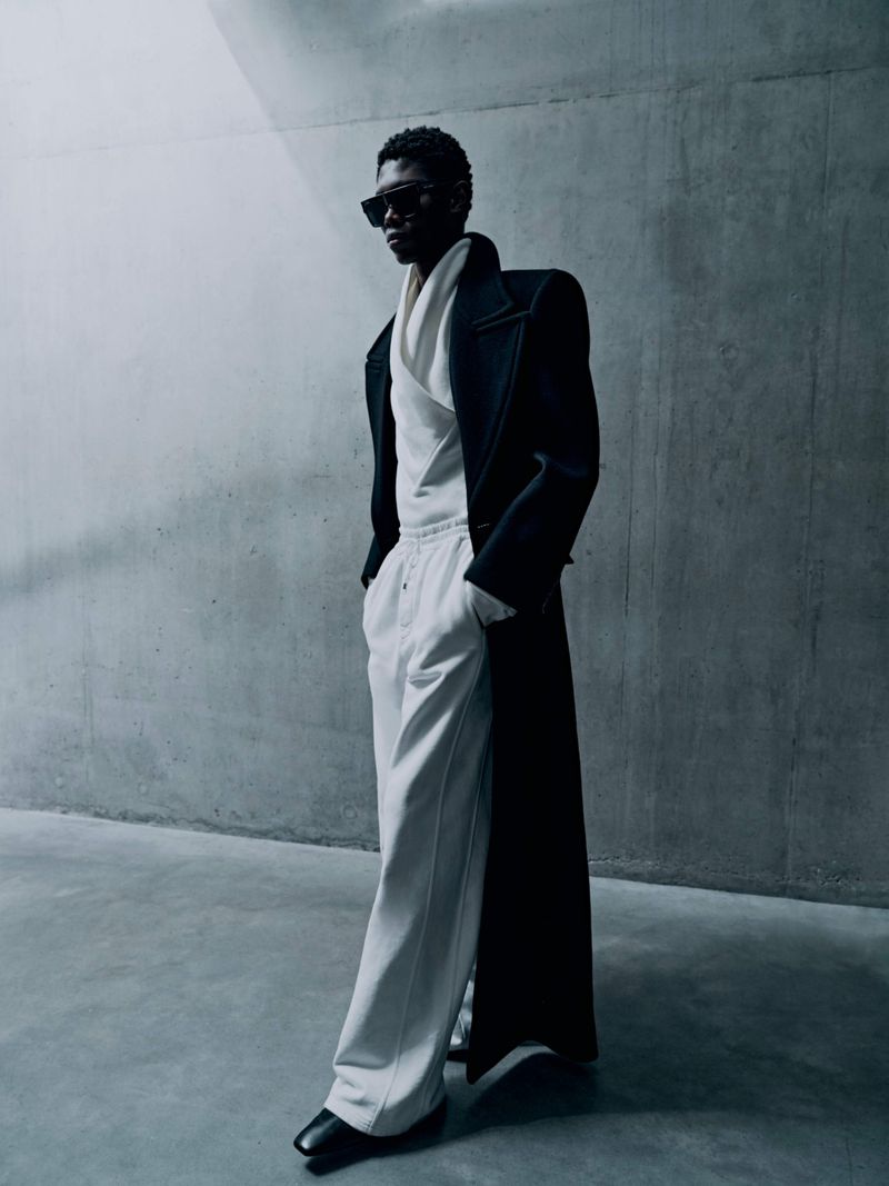 A slender, dark-skinned male fashion model with sunglasses poses in an oversized black coat and white pants against a grey wall.