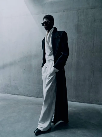 A slender, dark-skinned male fashion model with sunglasses poses in an oversized black coat and white pants against a grey wall.
