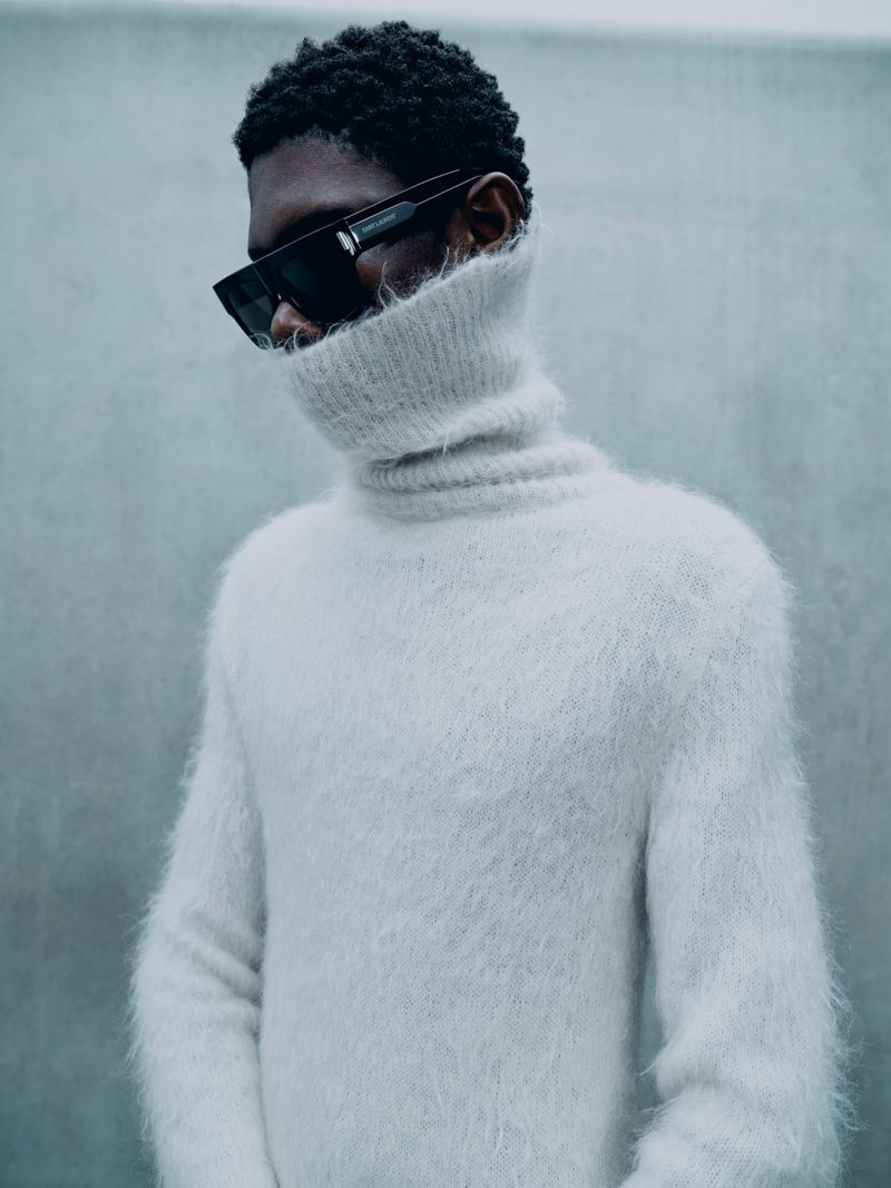 Close-up of a black man with a high-necked sweater and sunglasses in a studio setting against a light gray backdrop, fashion photography.