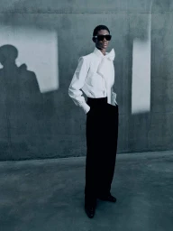 Model in black sunglasses wearing a white bow blouse and black trousers against a concrete wall