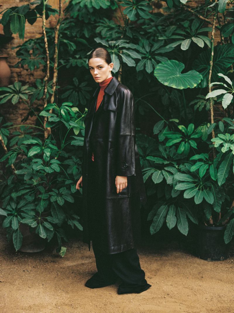 Elegant adult woman in a black leather coat and red turtleneck poses amid lush green plants and a brick wall, captured in soft light.