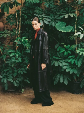 Elegant adult woman in a black leather coat and red turtleneck poses amid lush green plants and a brick wall, captured in soft light.