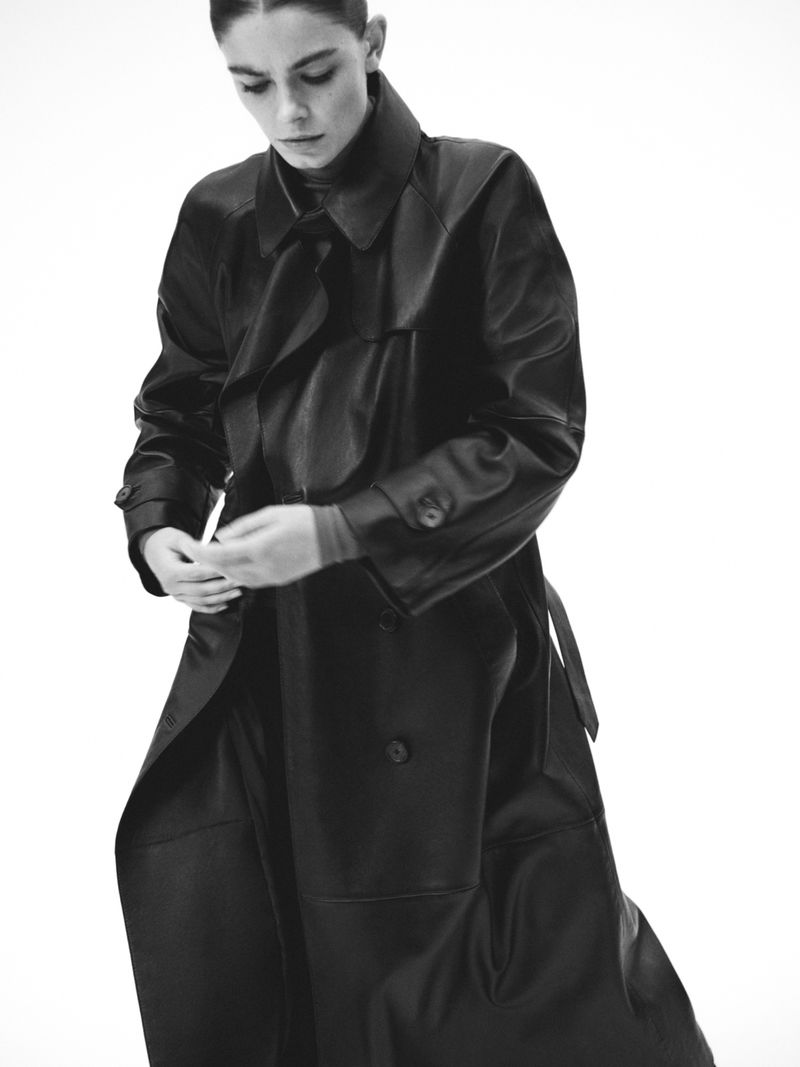 Monochrome portrait of a woman in a dark, oversized coat, buttoning it, in a fashion studio with white background.