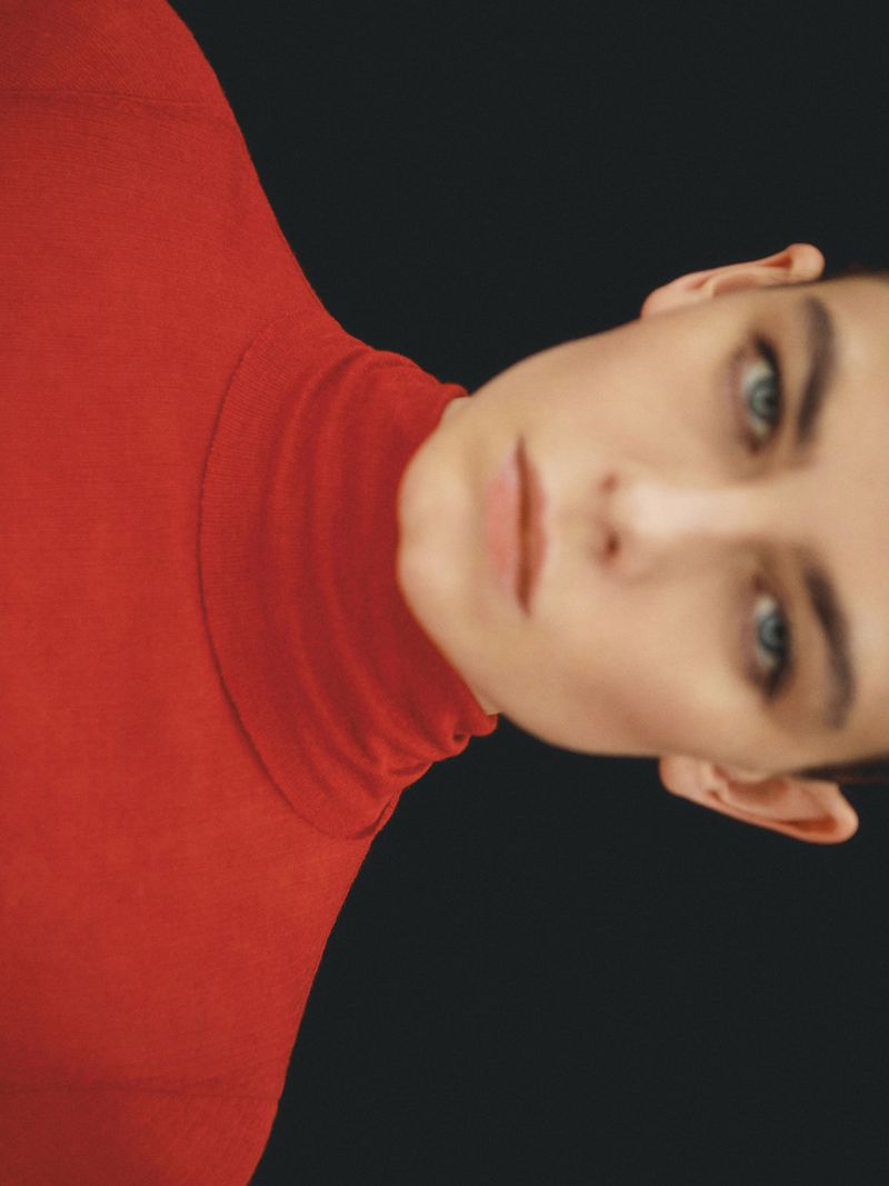 Close-up portrait of a young adult in a red turtleneck against a solid black backdrop, capturing the model's neutral expression and blue eyes.