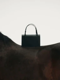 A structured dark leather handbag resting on the back of a dark horse against a plain white background.