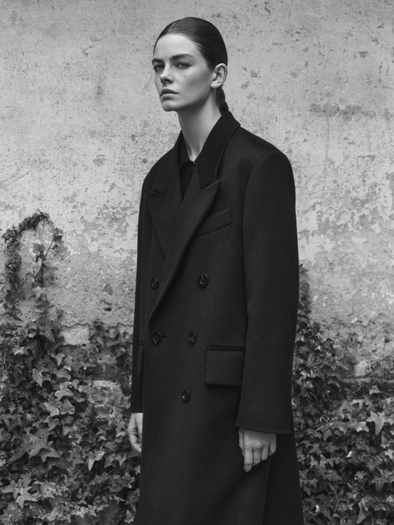 Model wearing a dark double-breasted coat standing against a weathered stone wall with climbing vines