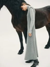 Model in grey turtleneck maxi dress leaning against a dark horse on white