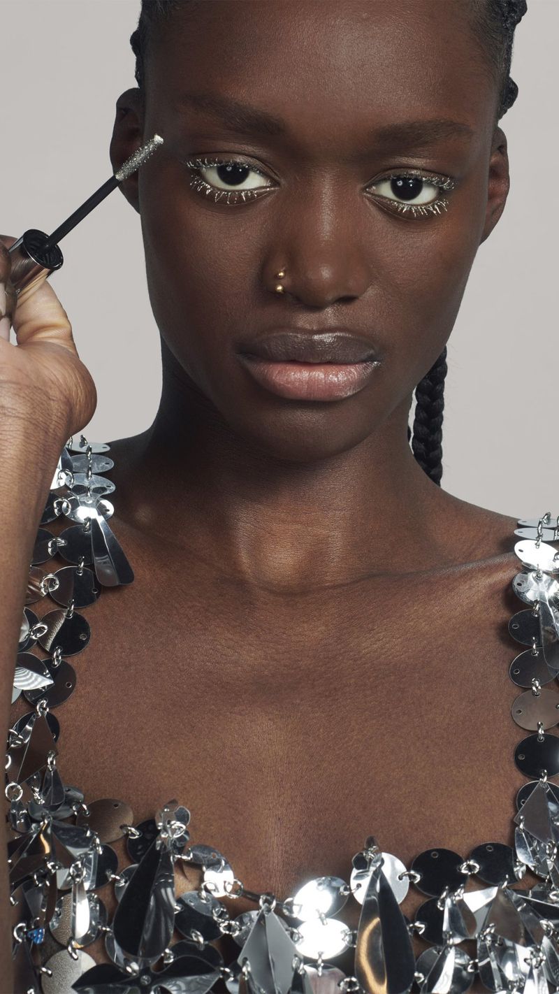 Close-up shot of an African American female model having silver cosmetic glitter applied to her eyelashes by an unseen person.