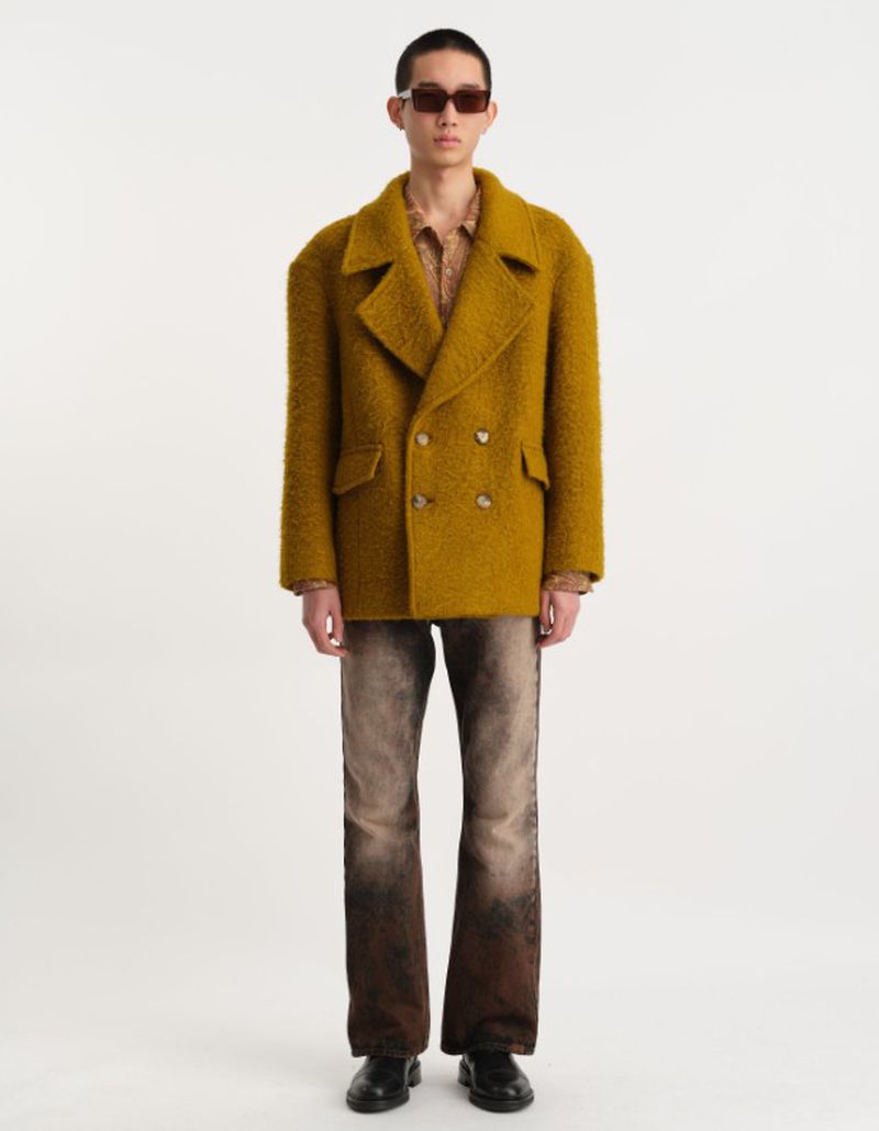 A young adult man stands in a light studio wearing a mustard-colored wool double-breasted coat with brown jeans and black shoes.