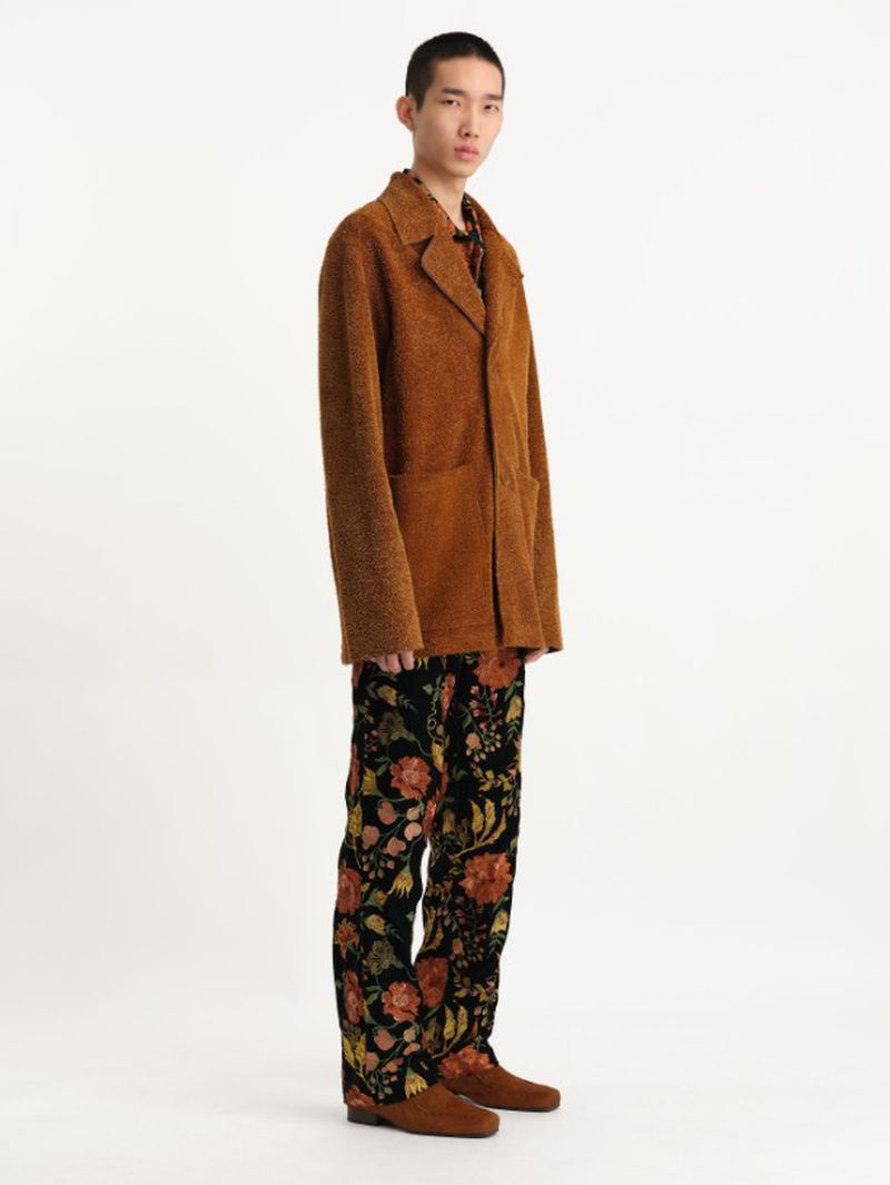Profile of an Asian male model wearing a brown textured jacket and floral patterned pants against a white backdrop in a fashion studio.