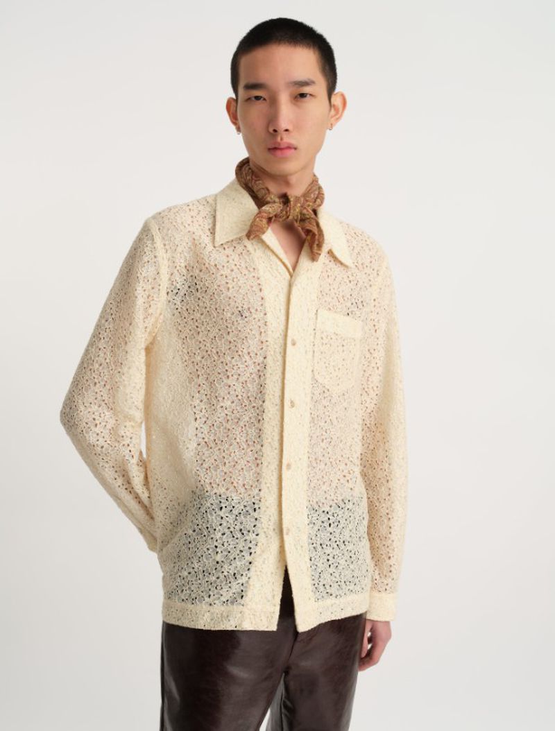 Man in a cream-colored lace shirt with brown leather pants and paisley scarf, posing indoors against a plain white backdrop.