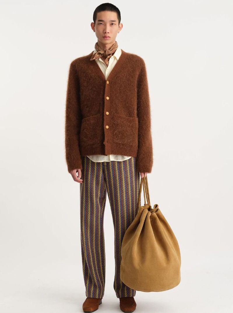 A slender man in a brown cardigan, patterned trousers, and brown shoes stands in a photo studio holding a tan drawstring bag.