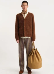 Male model wearing a brown cardigan, patterned trousers, and holding a large beige bag against a white background