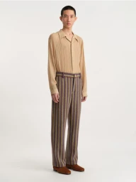 Male model wearing a pleated tan shirt and patterned trousers against a neutral white seamless studio background.