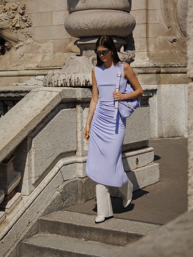 A woman in a periwinkle dress and white boots descends stone stairs in front of a stone monument on a sunny day.