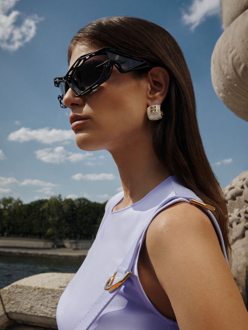 Woman in lilac dress and modern black sunglasses gazes to the side near a stone structure and river under a blue sky with light clouds.