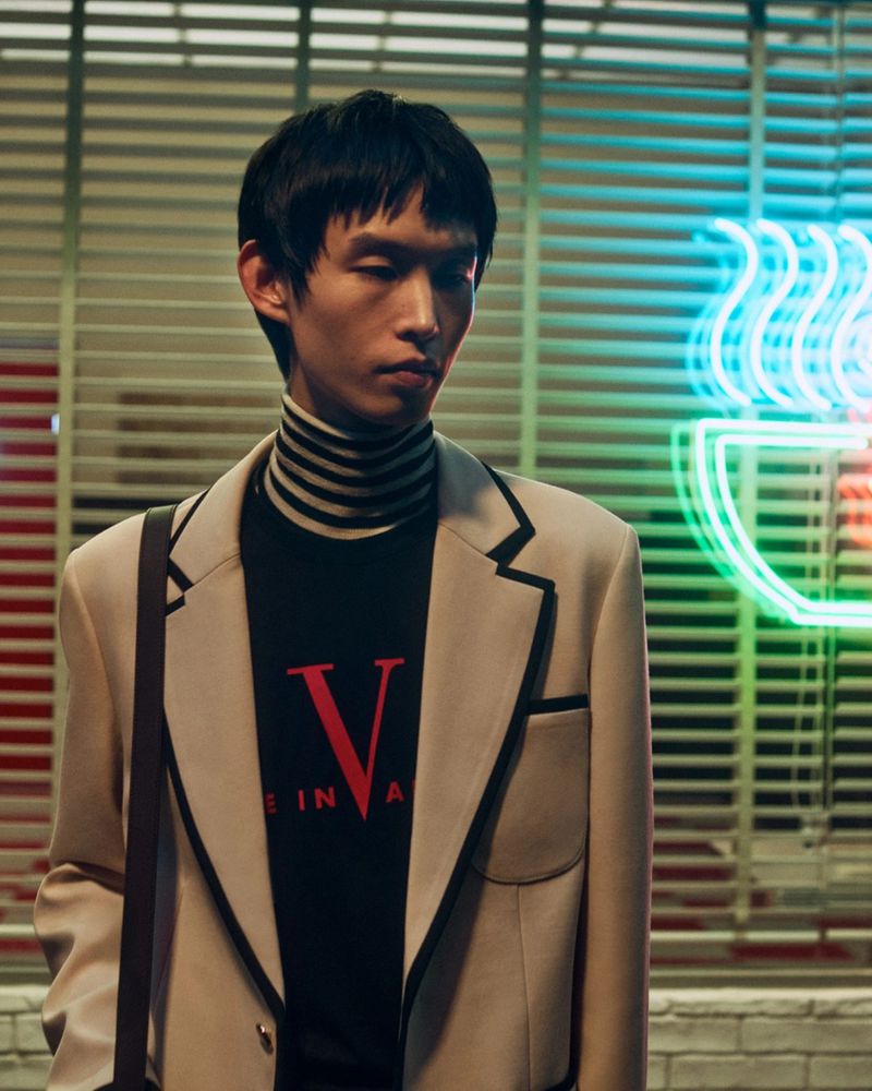 An Asian male model wearing a black and beige blazer and striped turtleneck stands in front of a glowing neon sign and blinds.