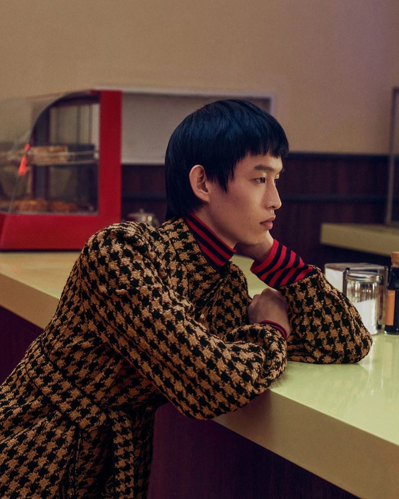 Asian male fashion model poses indoors in a houndstooth overcoat with a red striped turtleneck sweater with a diner counter in the background.