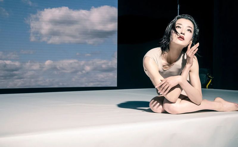 An adult woman poses in front of a light blue wall with clouds; she's kneeling with crossed arms, and is well lit by the sun.