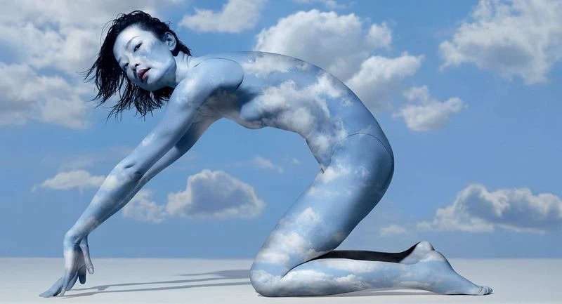 Woman kneeling with body paint of cloudy blue sky, body art and artistic photography in blue and white tones, serene and conceptual.