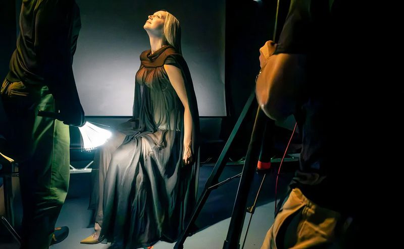 Woman posing under studio lights while seated in a dark gown. Crew members stand nearby with lighting equipment.