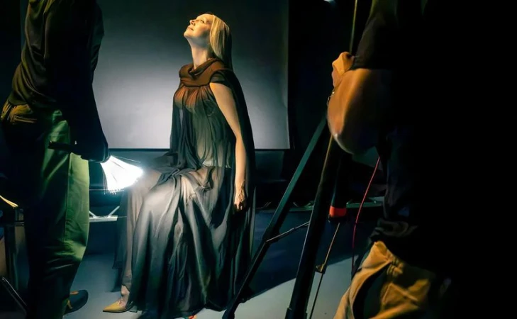 Woman posing under studio lights while seated in a dark gown. Crew members stand nearby with lighting equipment.