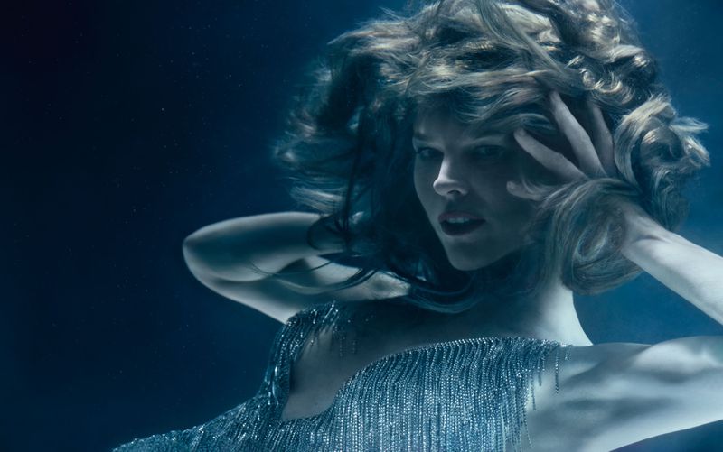 Underwater portrait of an adult female with blonde hair, wearing a dress with metallic fringe, shot in cool, low lighting.