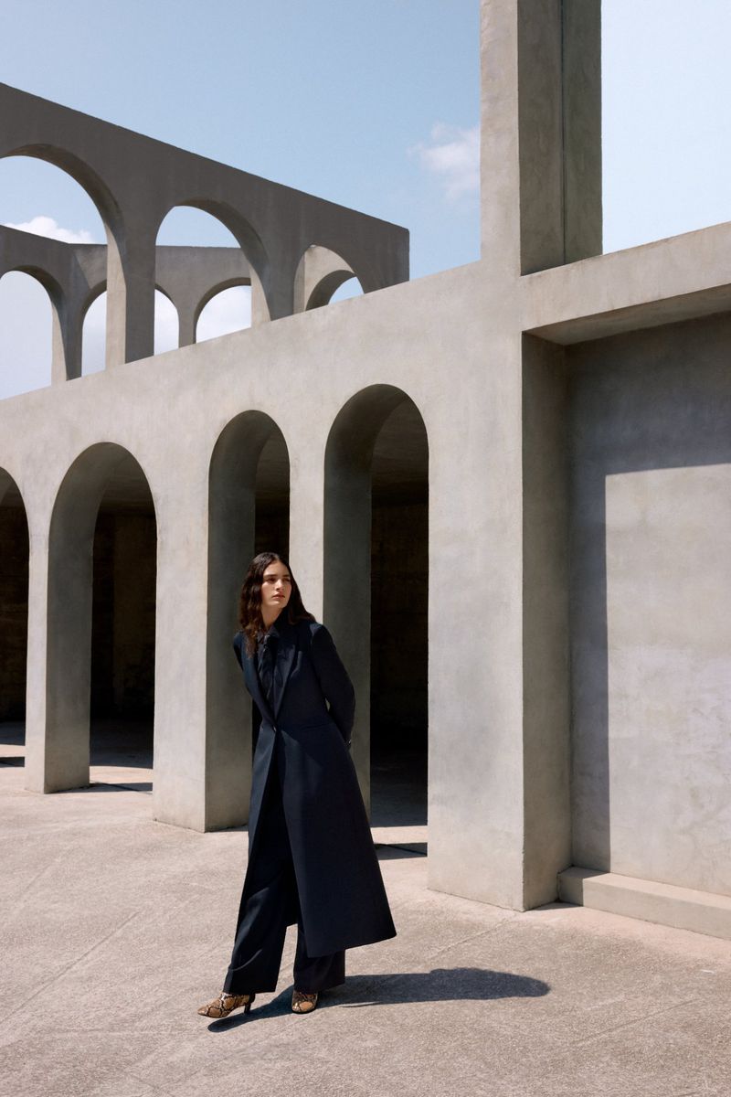 Woman in a long dark coat and wide-leg pants stands in front of a modern concrete building with arches on a sunny day.
