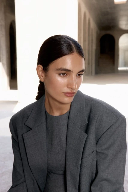 Portrait of adult female model in gray blazer and dress, posing in bright light near arched openings, neutral colors. Fashion, beauty.