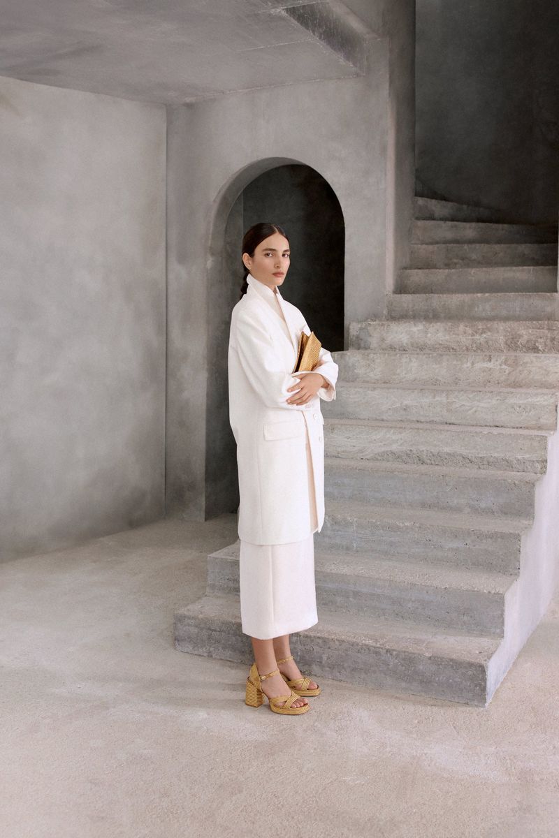 A woman stands indoors in a white coat and skirt, holding a book, near gray concrete stairs, with an arched opening in the background.