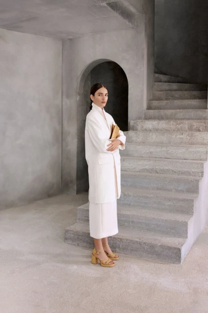 A woman stands indoors in a white coat and skirt, holding a book, near gray concrete stairs, with an arched opening in the background.