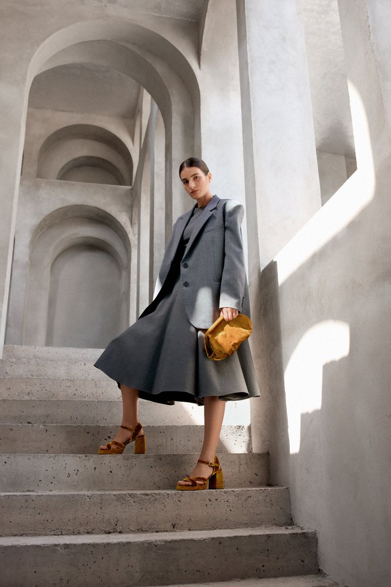 Adult woman in grey suit and heels poses on concrete stairs, holding a yellow purse with arched passageway in the background.