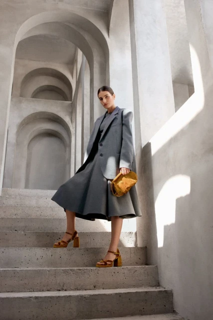 Adult woman in grey suit and heels poses on concrete stairs, holding a yellow purse with arched passageway in the background.