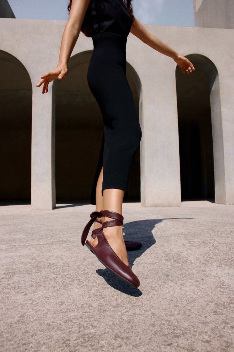 Woman in black dress and maroon ballet flats poses near a concrete wall with archways in natural light, showcasing fashion and style.