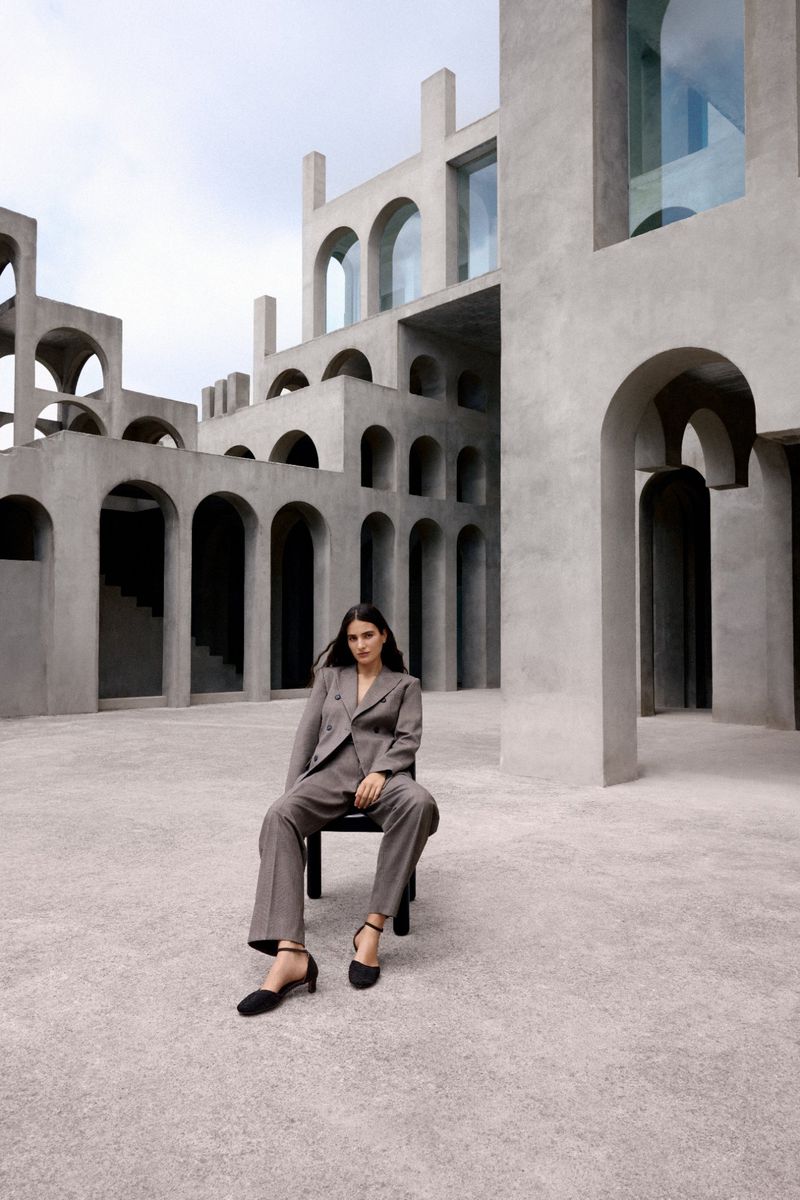 A woman in a gray suit sits on a black chair in front of an imposing architectural building with arches and open space.