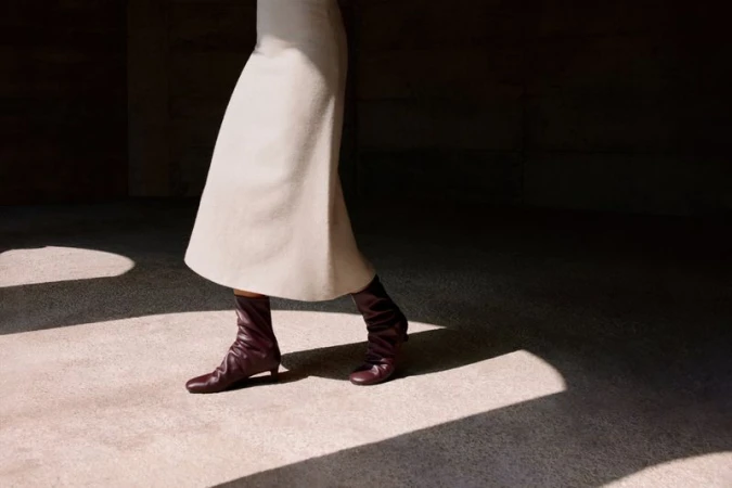 A fashion shot of a woman walking indoors, wearing a cream skirt and maroon ankle boots, with natural light casting shadows on the floor.