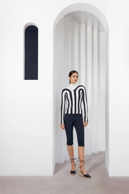 Woman in black and white striped top and dark capri pants stands in white arched doorway with black shoes with ankle ties.