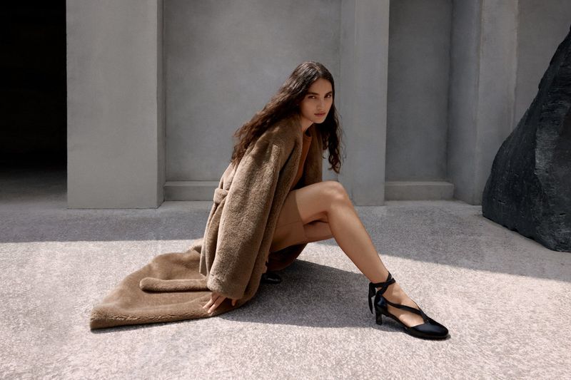 A brunette woman in a tan coat and dress poses in black shoes in front of a gray stone wall and black boulder formation.