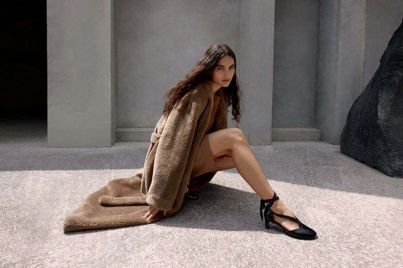 A brunette woman in a tan coat and dress poses in black shoes in front of a gray stone wall and black boulder formation.